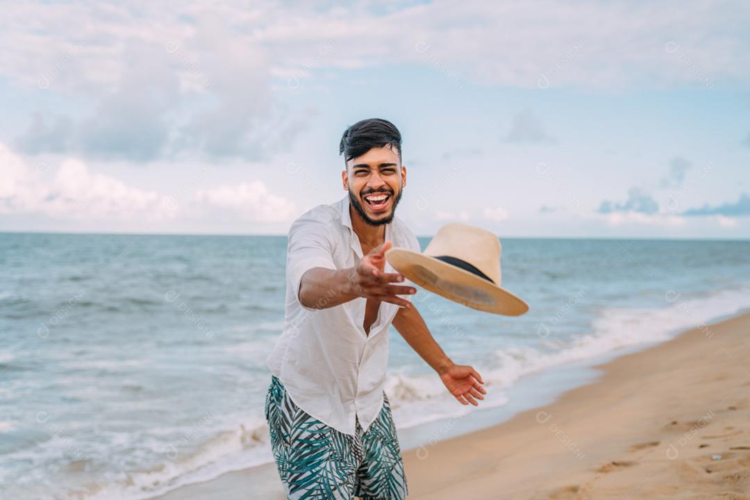 Homem latino-americano sorrindo e jogando seu chapéu olhando para a câmera na praia em um lindo dia de verão