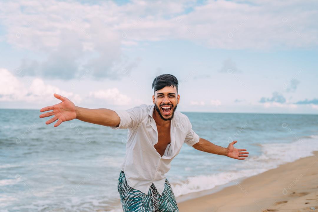 Homem latino-americano sorrindo e jogando seu chapéu olhando para a câmera na praia em um lindo dia de verão