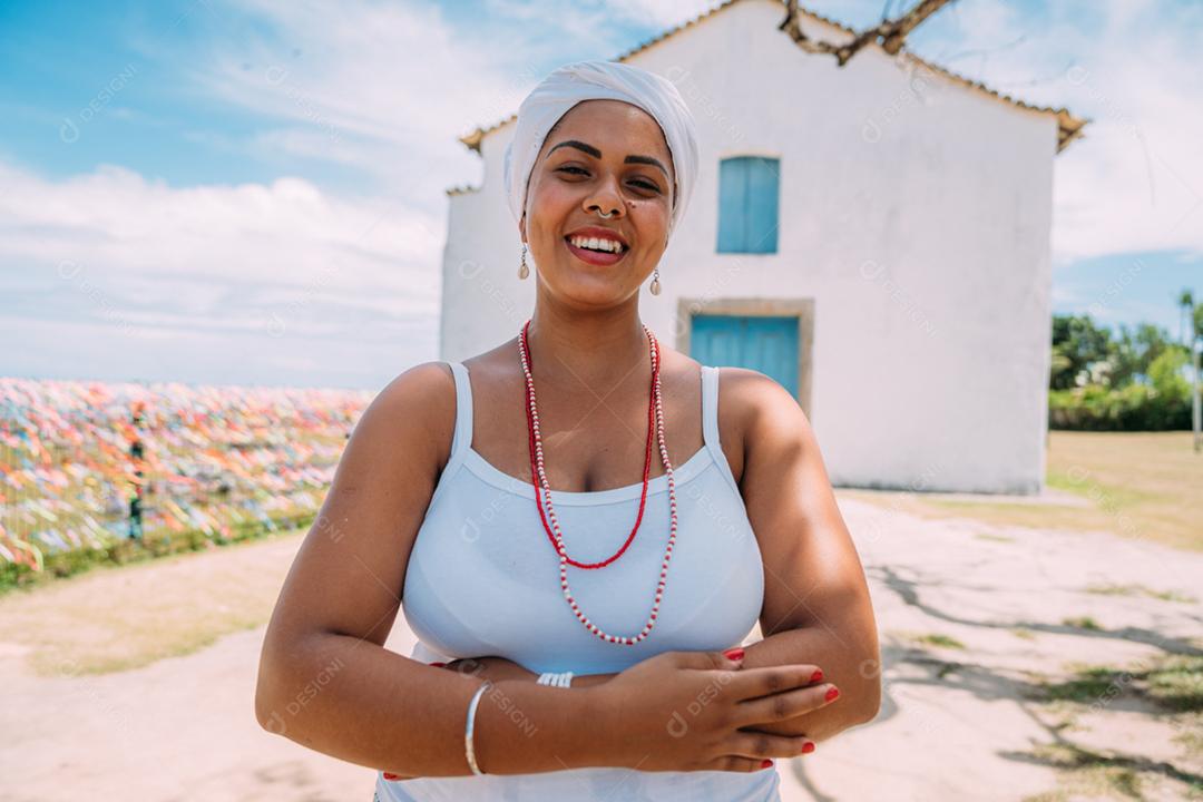 Feliz mulher brasileira vestida com traje tradicional baiano, braços cruzados no centro histórico de Porto Seguro ao fundo