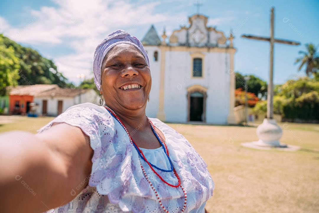 Feliz mulher brasileira de ascendência africana vestida com o tradicional vestido baiano fazendo uma selfie em frente à igreja