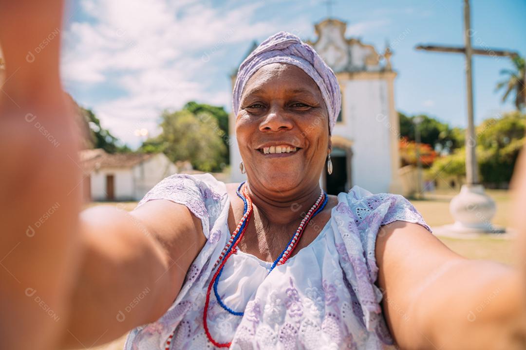 Feliz mulher brasileira de ascendência africana vestida com o tradicional vestido baiano fazendo uma selfie em frente à igreja