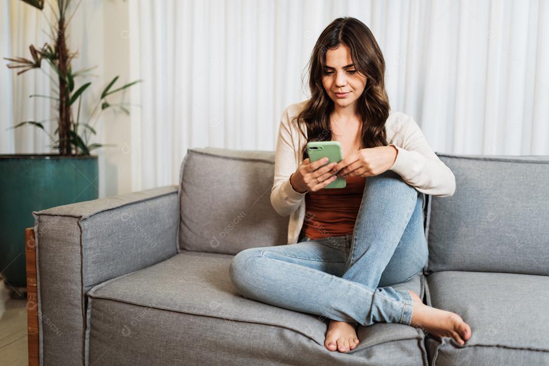 Feliz dama latina relaxa em casa sozinha, sente-se na sala e compartilhe boas notícias nas mídias sociais via celular