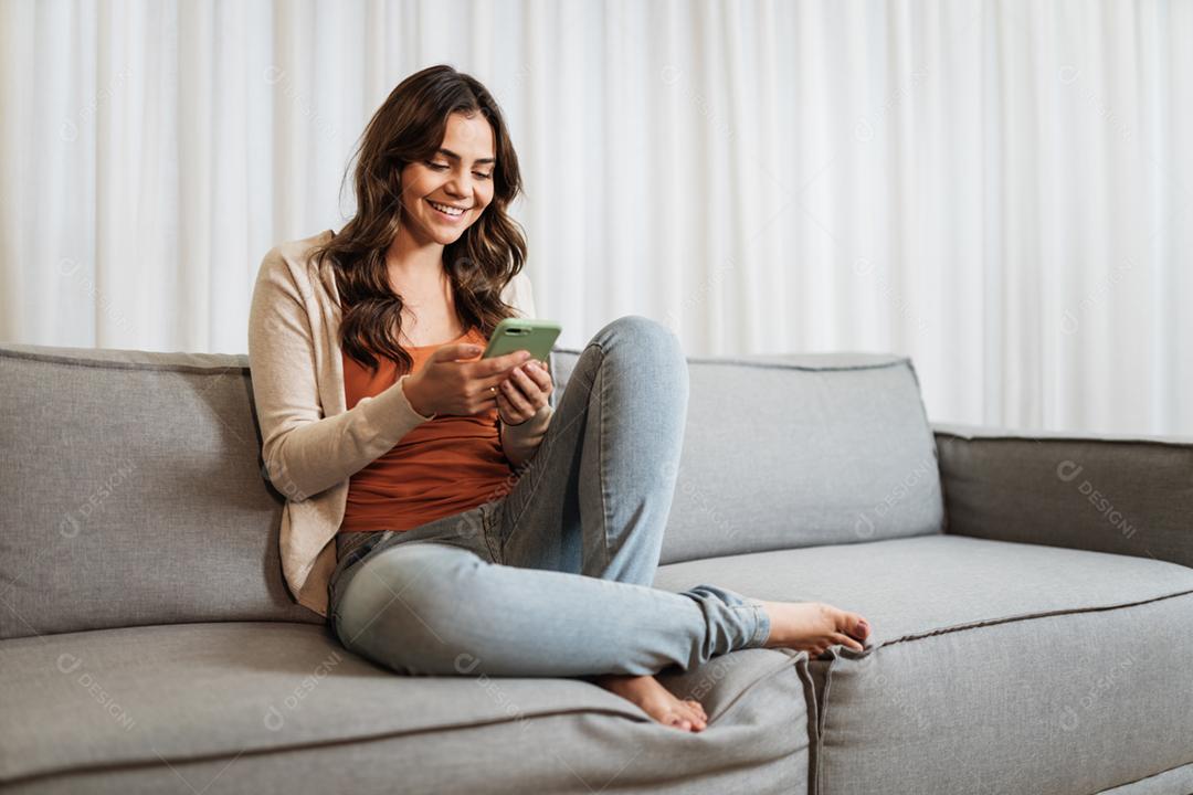 Feliz dama latina relaxa em casa sozinha, sente-se na sala e compartilhe boas notícias nas mídias sociais via celular