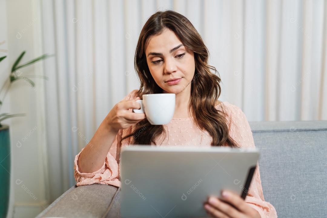A senhora latina feliz relaxa tomando café em casa sozinha sentada na sala usando tablet digital.