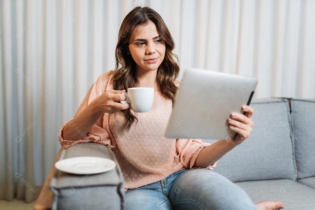 A senhora latina feliz relaxa tomando café em casa sozinha sentada na sala usando tablet digital.