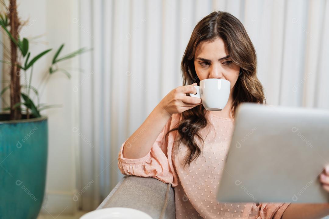 A senhora latina feliz relaxa tomando café em casa sozinha sentada na sala usando tablet digital.