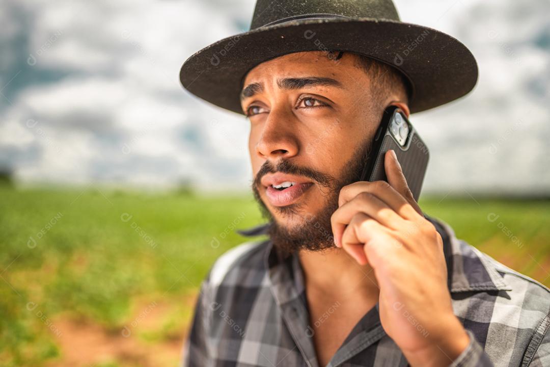 Agricultor latino-americano trabalhando na plantação. Jovem falando no celular, usando chapéu