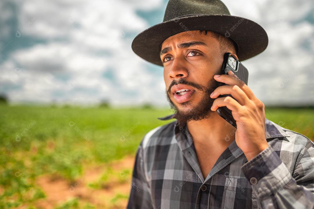 Agricultor latino-americano trabalhando na plantação. Jovem falando no celular, usando chapéu