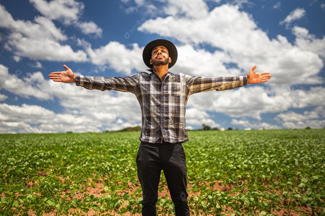Feliz jovem agricultor latino-americano desfrutando de liberdade com as mãos abertas na fazenda e no fundo do céu por do sol.