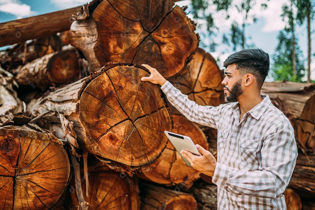 Retrato de jovem latino trabalhando em tablet ao lado de troncos de árvores.