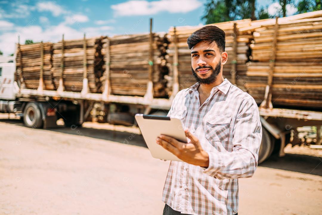 Retrato de jovem latino trabalhando com um tablet digital ao lado de troncos de árvores no caminhão.