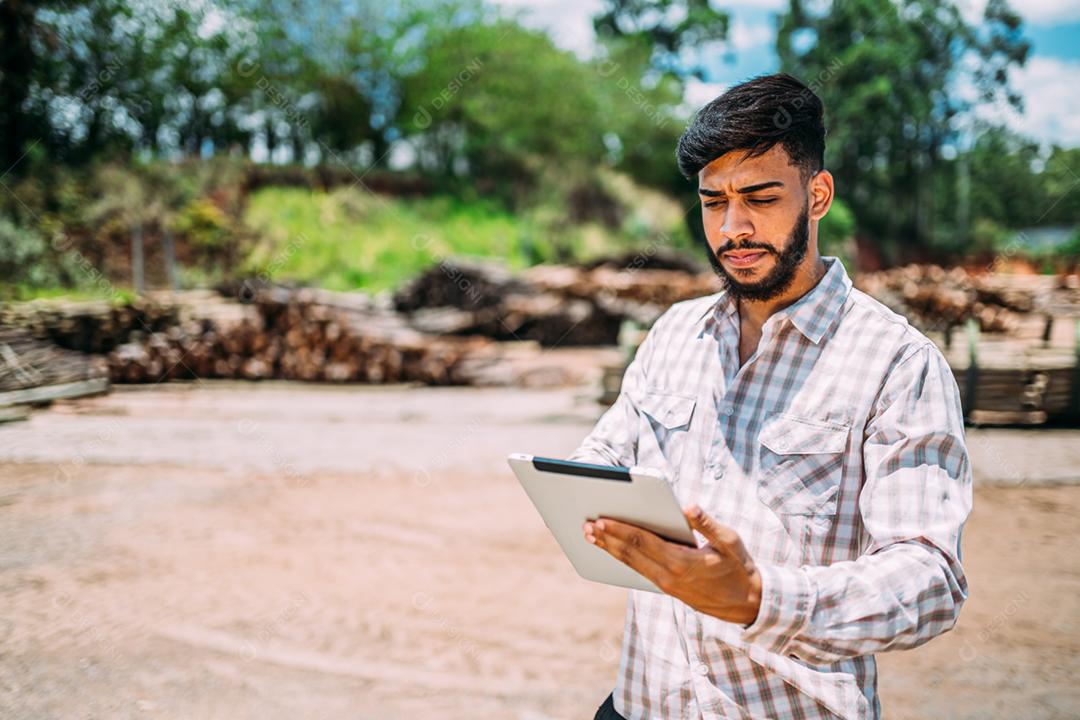 Retrato de jovem latino trabalhando em tablet ao lado de troncos de árvores.