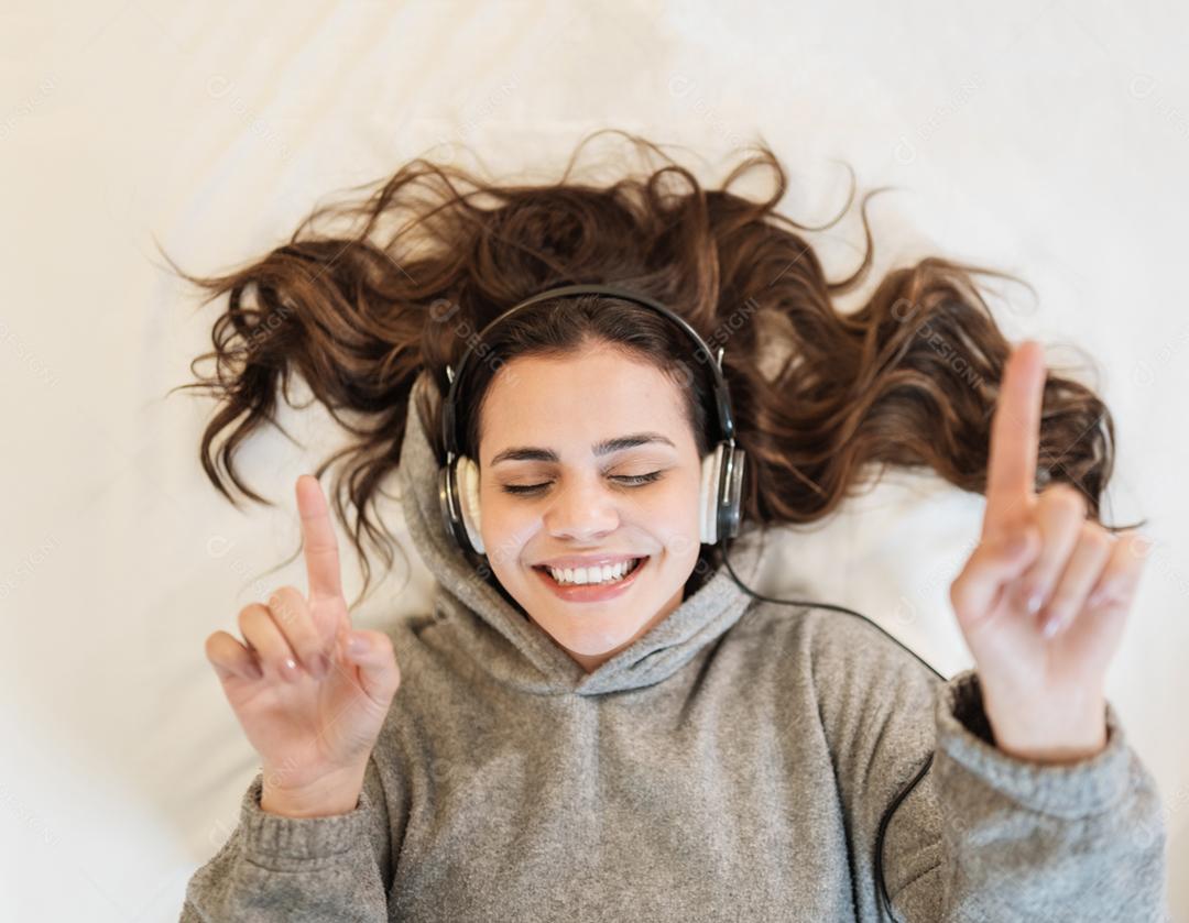 Mulher latina feliz usa fones de ouvido deitada na cama ouvindo música cantando com os olhos fechados