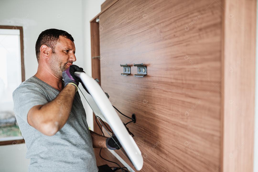 Conceito de serviço de entrega de móveis. Trabalhador latino de uniforme instalando televisão na parede dentro de casa.