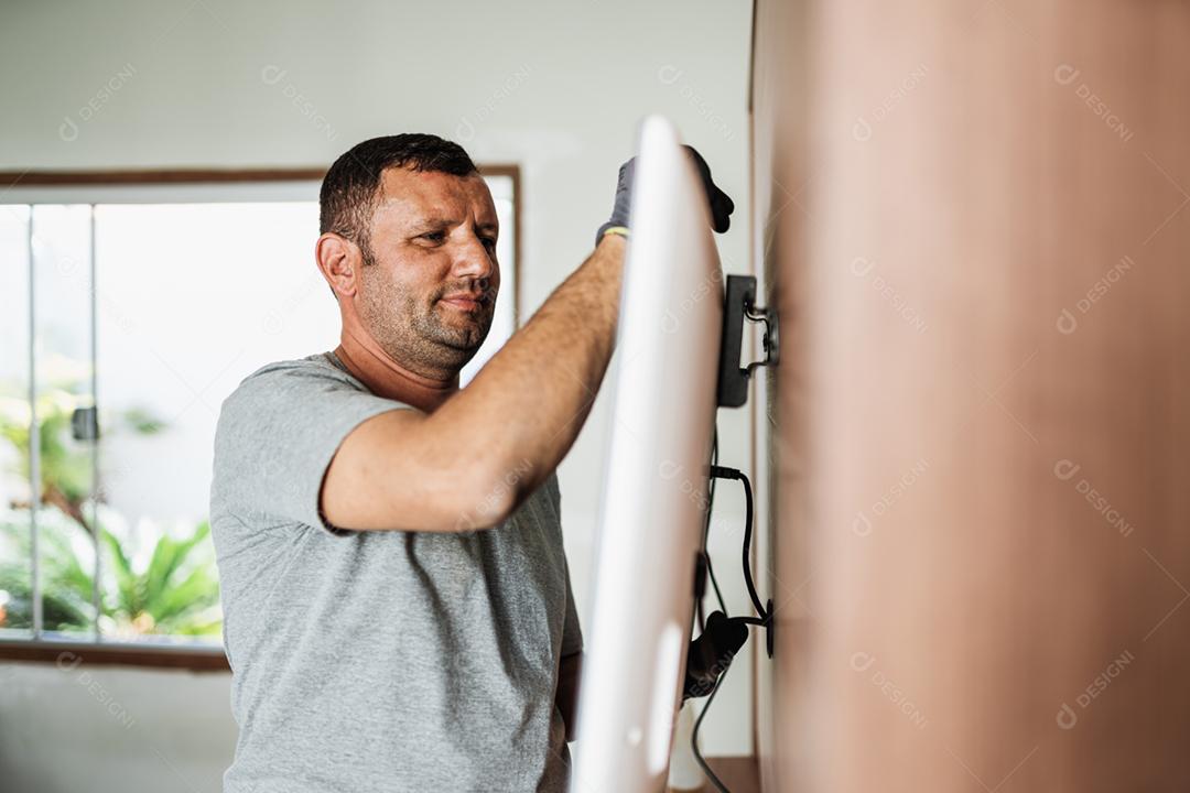 Furniture delivery service concept. Latin worker in uniform installing television on wall indoors.