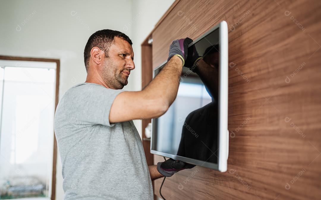 Conceito de serviço de entrega de móveis. Trabalhador latino de uniforme instalando televisão na parede dentro de casa.