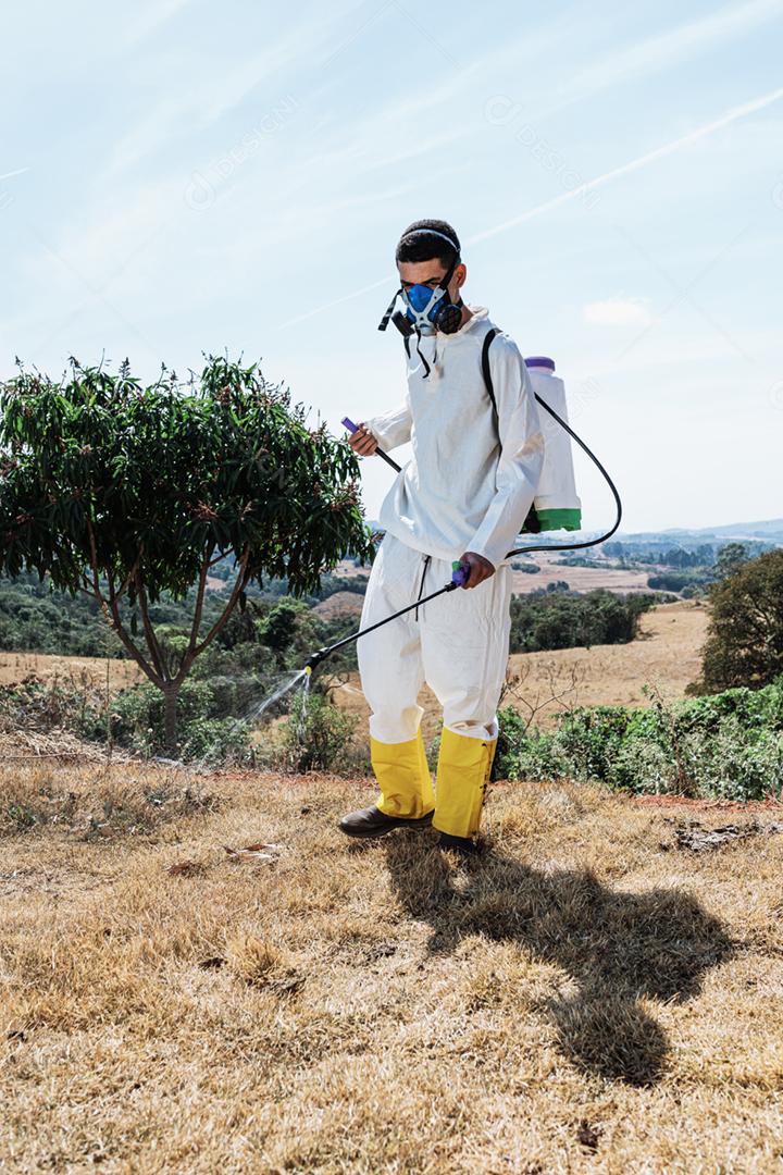 Homem latino vestindo roupas apropriadas e máscara protetora pulverizando ervas daninhas no terreno