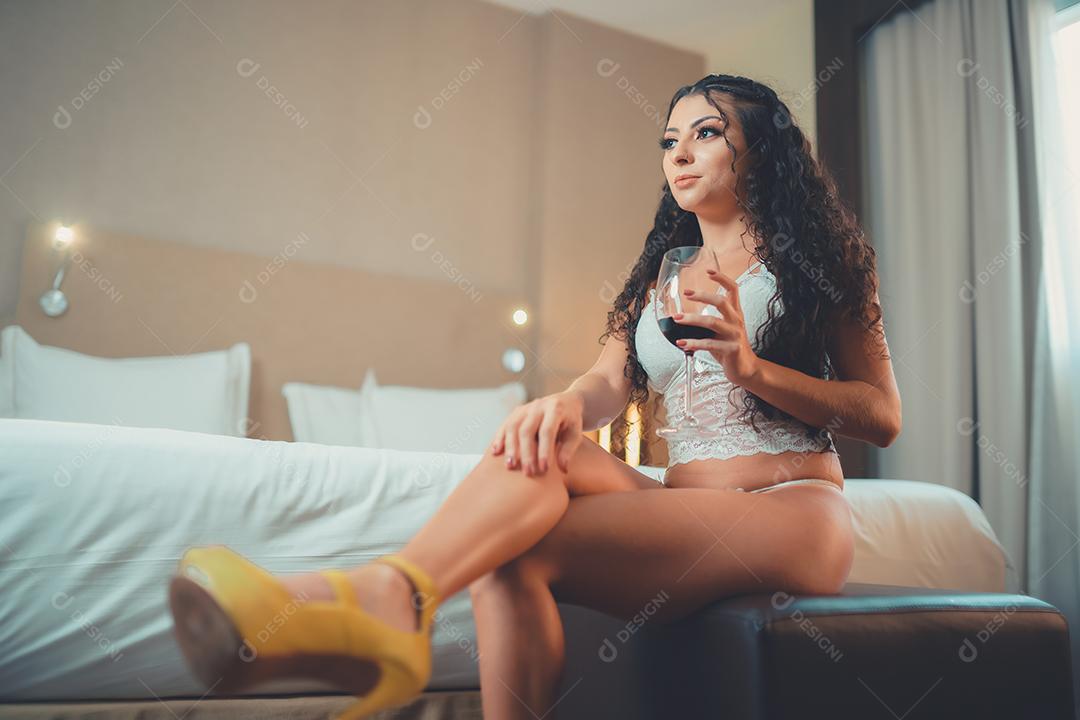 Mulher linda sexy sentada na cama com vinho no copo. Jovem cabelo encaracolado, elegante e bem sucedida. Bela latina