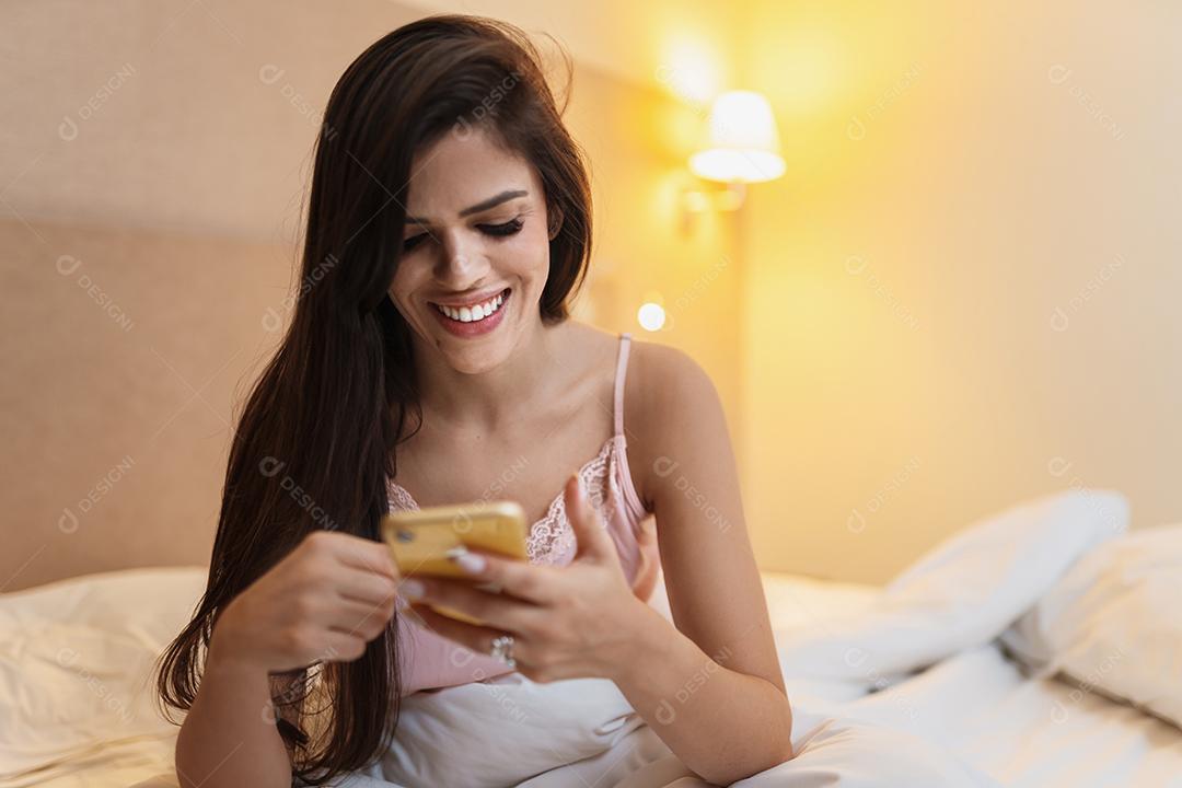 Linda jovem sorridente latina deitada na cama usando um telefone