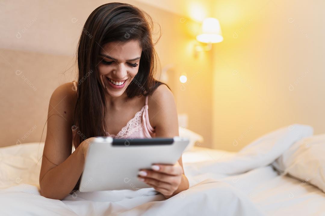 Bela jovem sorridente Latina deitada na cama usando um tablet digital no quarto dela.