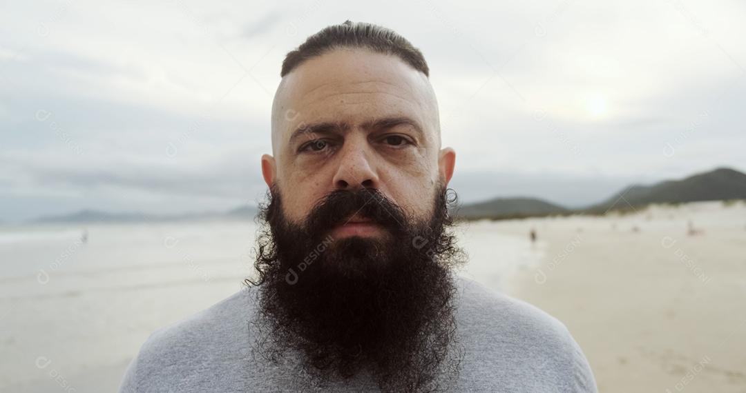 Homem latino sério com barba grande em uma praia olhando para a câmera.