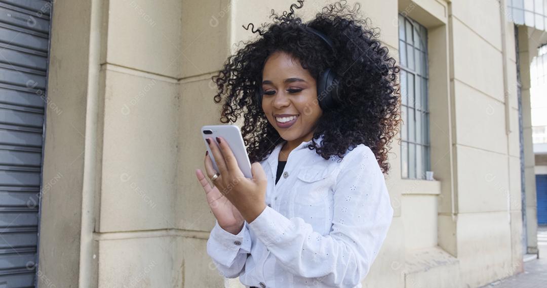 Jovem negra de cabelo encaracolado andando usando o celular. Mensagens de texto na rua. Cidade grande.