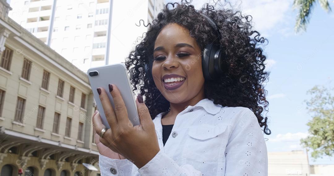 Jovem negra de cabelo encaracolado andando usando o celular. Mensagens de texto na rua. Cidade grande.