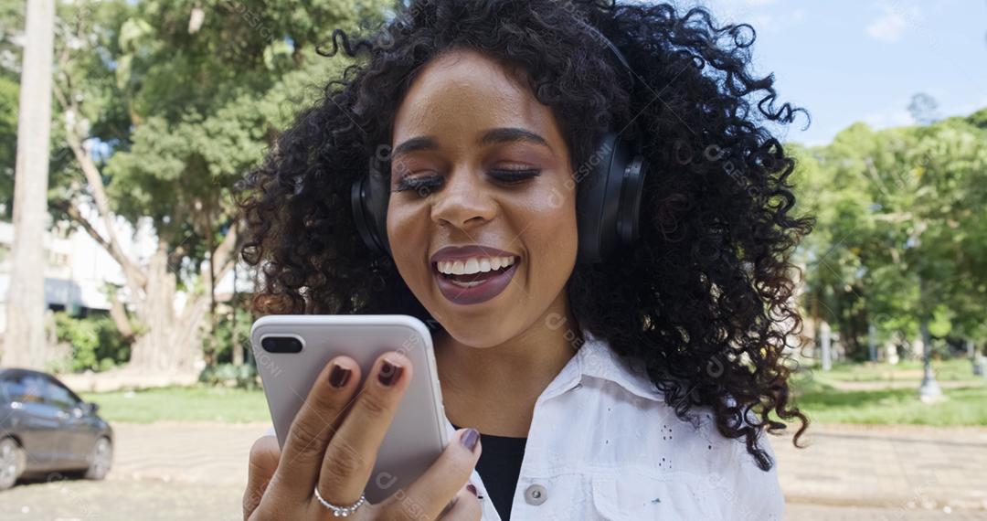 Jovem negra de cabelo encaracolado andando usando o celular. Mensagens de texto na rua. Cidade grande.