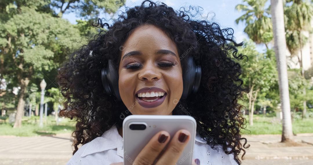 Jovem negra de cabelo encaracolado andando usando o celular. Mensagens de texto na rua. Cidade grande.