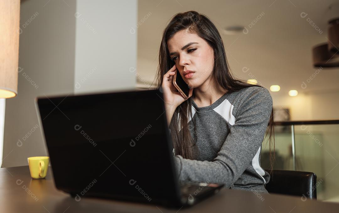 Jovem linda brasileira usando smartphone e trabalhando com laptop enquanto está sentado na mesa de escritório