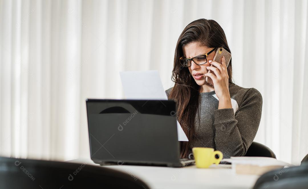 Jovem linda brasileira usando smartphone e trabalhando com laptop enquanto está sentado na mesa de escritório