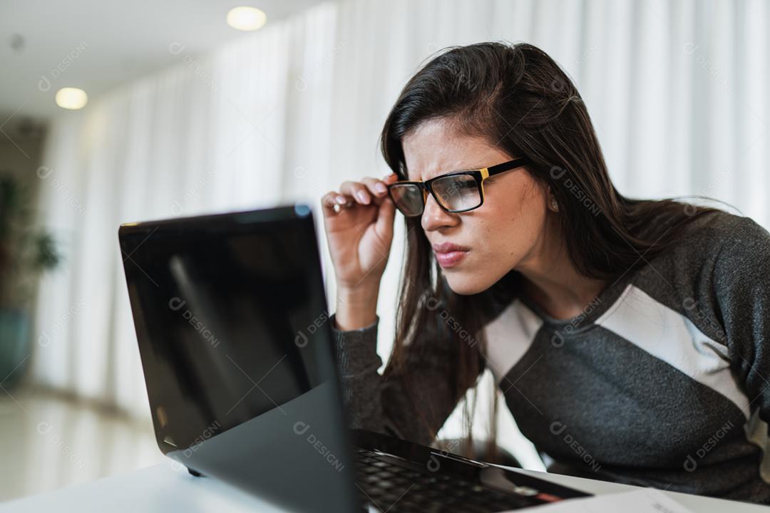 Mulher latina com problemas de visão tentando ler conteúdo online em um laptop em casa.