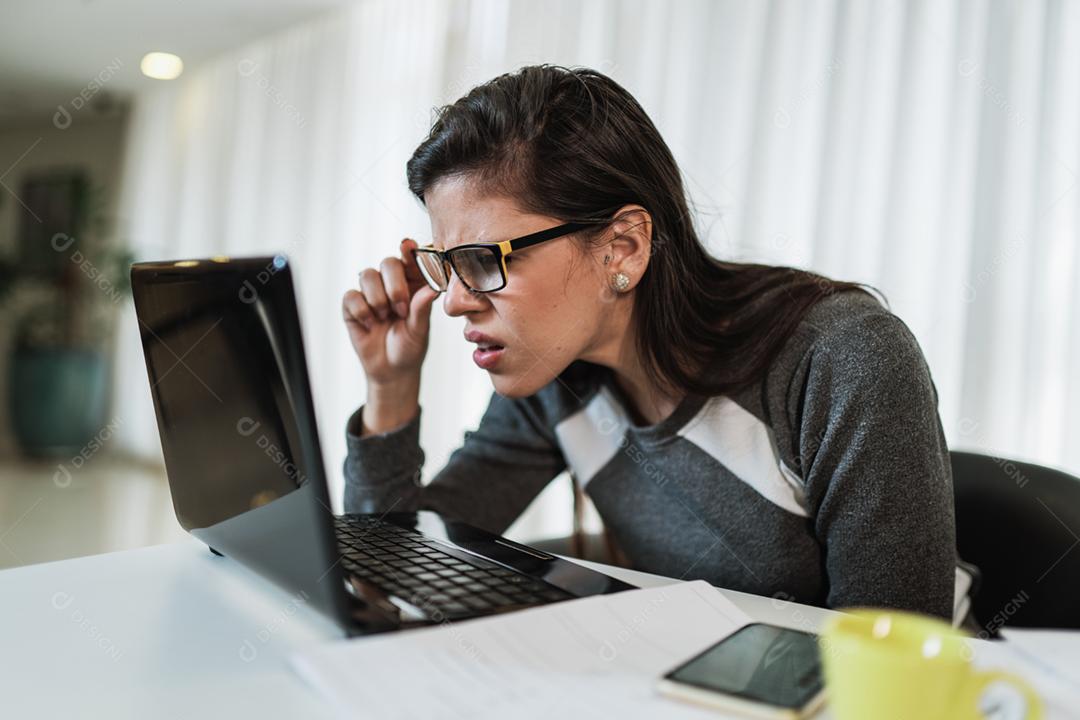 Mulher latina com problemas de visão tentando ler conteúdo online em um laptop em casa.