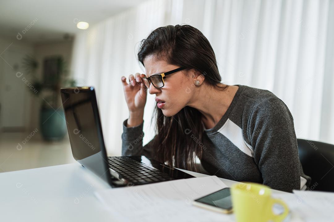 Mulher latina com problemas de visão tentando ler conteúdo online em um laptop em casa.