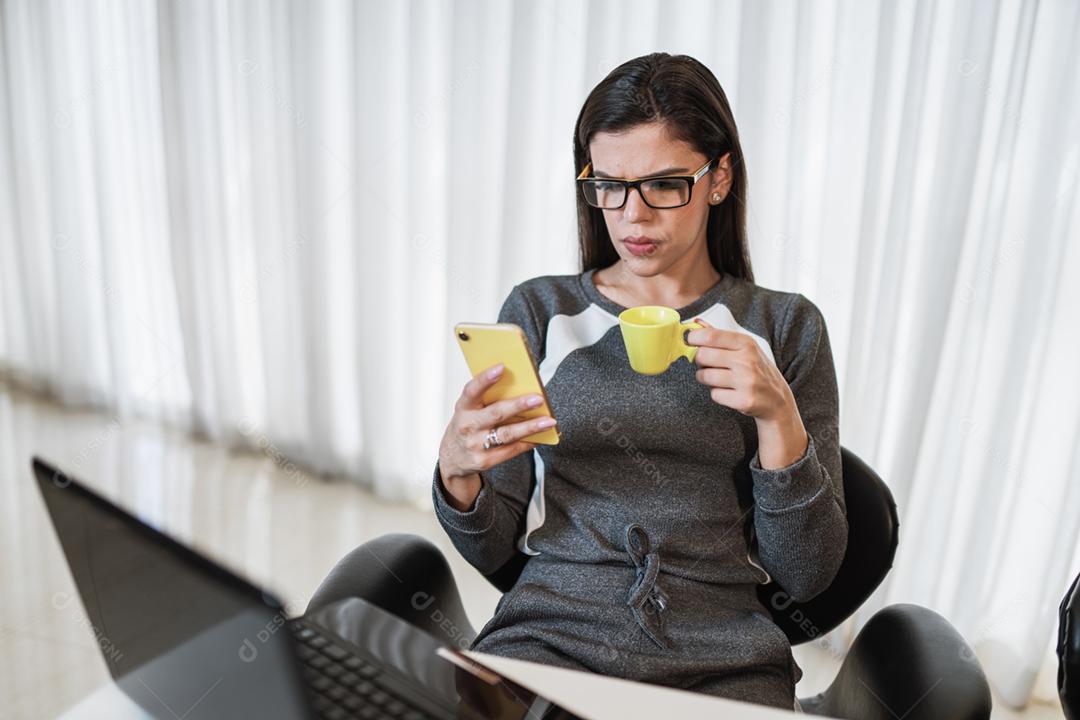 Jovem linda brasileira usando smartphone e trabalhando com laptop enquanto está sentado na mesa de escritório
