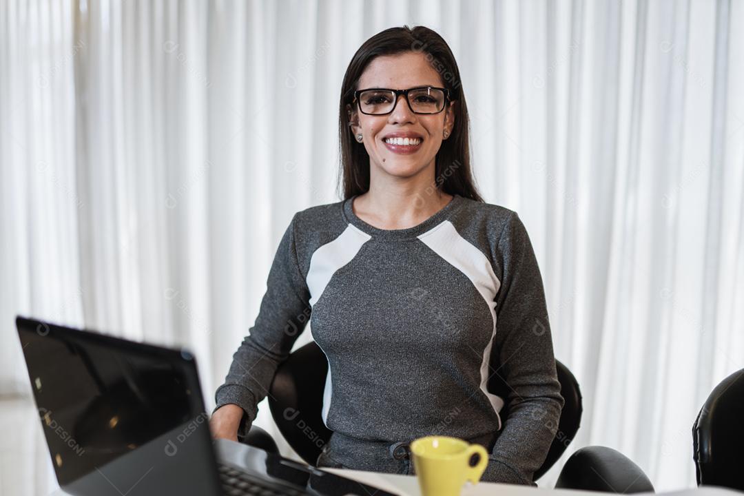 Jovem linda brasileira usando smartphone e trabalhando com laptop enquanto está sentado na mesa de escritório