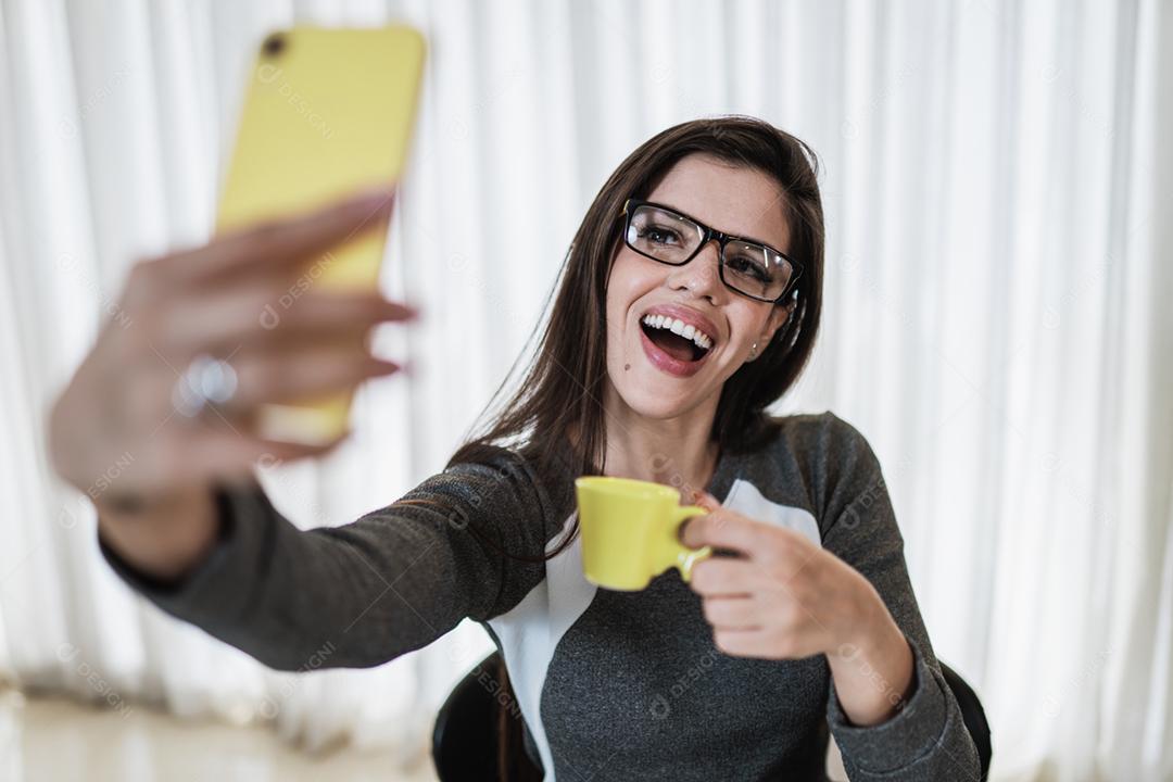 Retrato de uma alegre garota latina vestida com roupas cinzentas segurando uma xícara de café amarela enquanto tira uma selfie em casa
