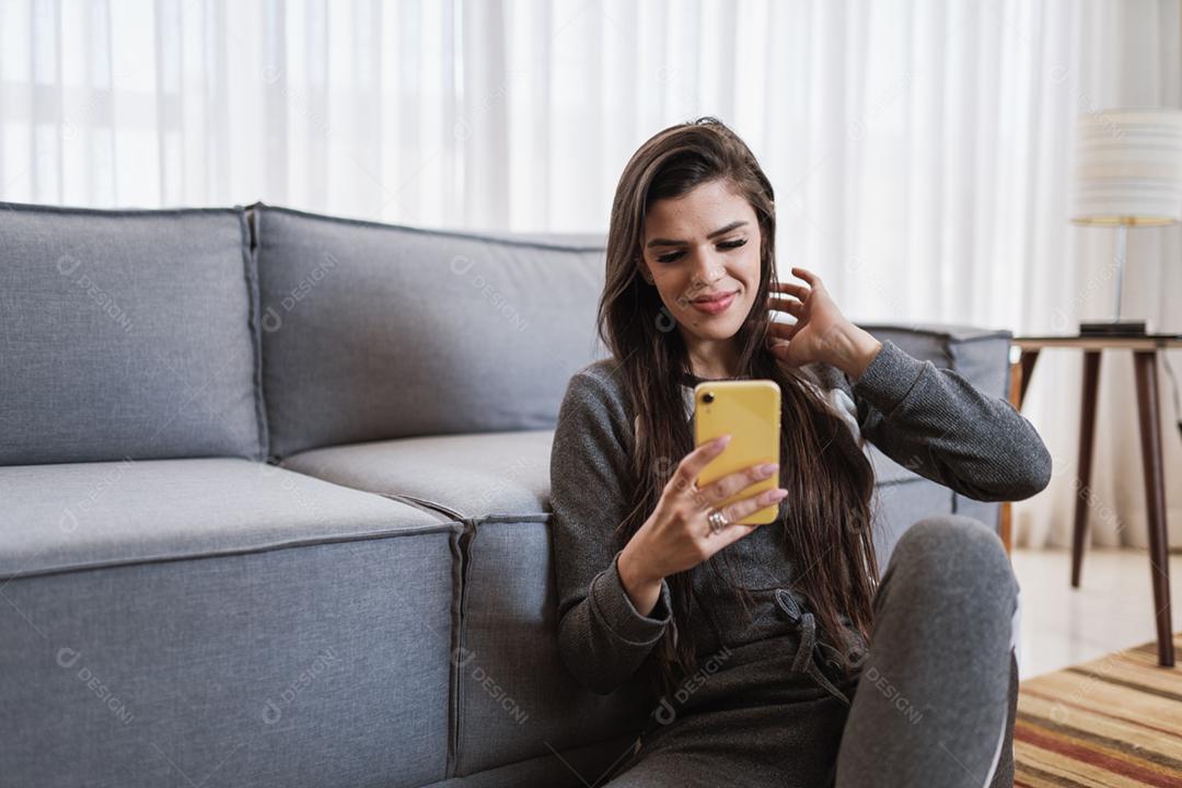 Feliz dama latina relaxa em casa sozinha, sente-se na sala e compartilhe boas notícias nas mídias sociais via celular.