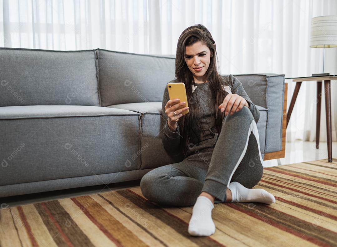 Feliz dama latina relaxa em casa sozinha, sente-se na sala e compartilhe boas notícias nas mídias sociais via celular.