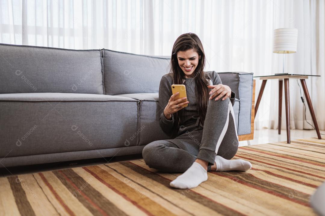 Feliz dama latina relaxa em casa sozinha, sente-se na sala e compartilhe boas notícias nas mídias sociais via celular.