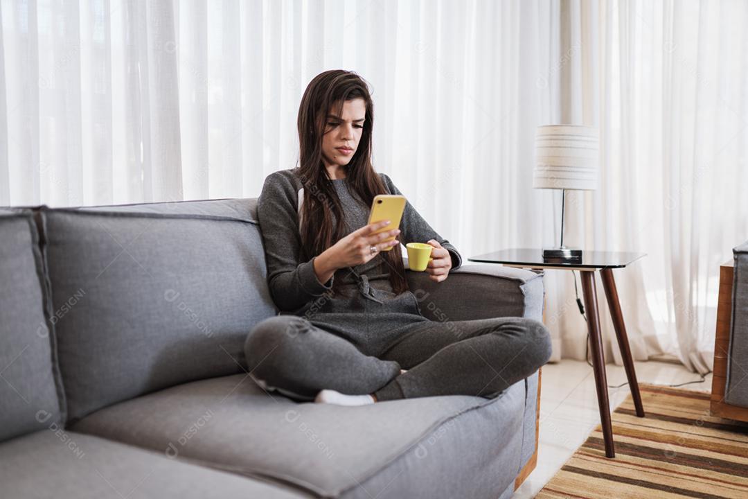 Feliz dama latina relaxa em casa sozinha, sente-se na sala e compartilhe boas notícias nas mídias sociais via celular.