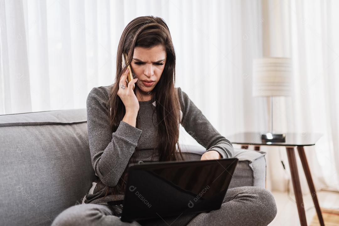 Jovem linda brasileira usando smartphone e trabalhando com laptop enquanto está sentado no sofá, trabalhando em casa conceito.