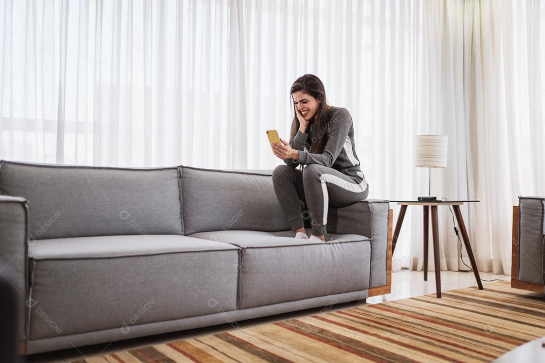 Feliz dama latina relaxa em casa sozinha, sente-se na sala e compartilhe boas notícias nas mídias sociais via celular