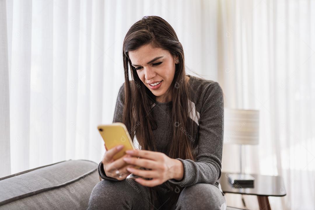 Feliz dama latina relaxa em casa sozinha, sente-se na sala e compartilhe boas notícias nas mídias sociais via celular