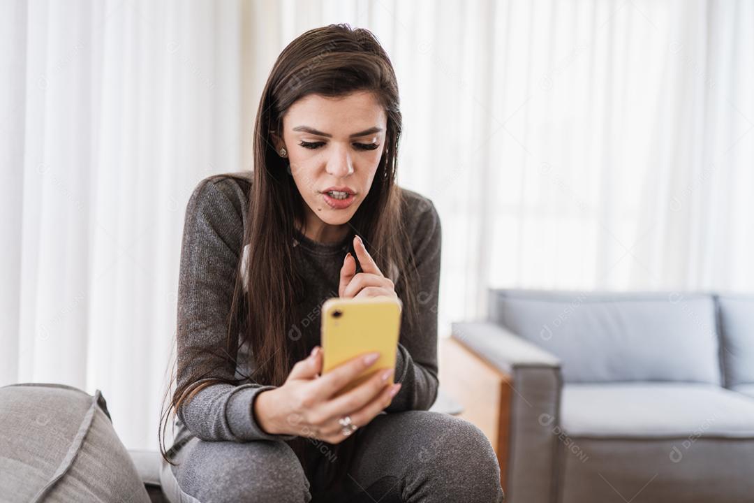 Feliz dama latina relaxa em casa sozinha, sente-se na sala e compartilhe boas notícias nas mídias sociais via celular