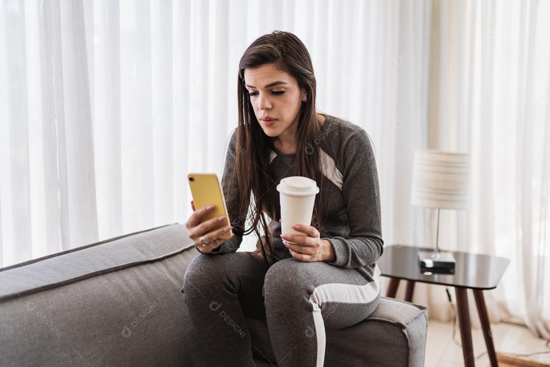 A senhora latina feliz relaxa tomando café em casa sozinha, sentada na sala, compartilha boas notícias nas mídias sociais via celular