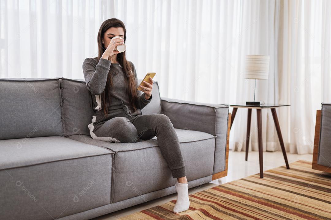 A senhora latina feliz relaxa tomando café em casa sozinha, sentada na sala, compartilha boas notícias nas mídias sociais via celular