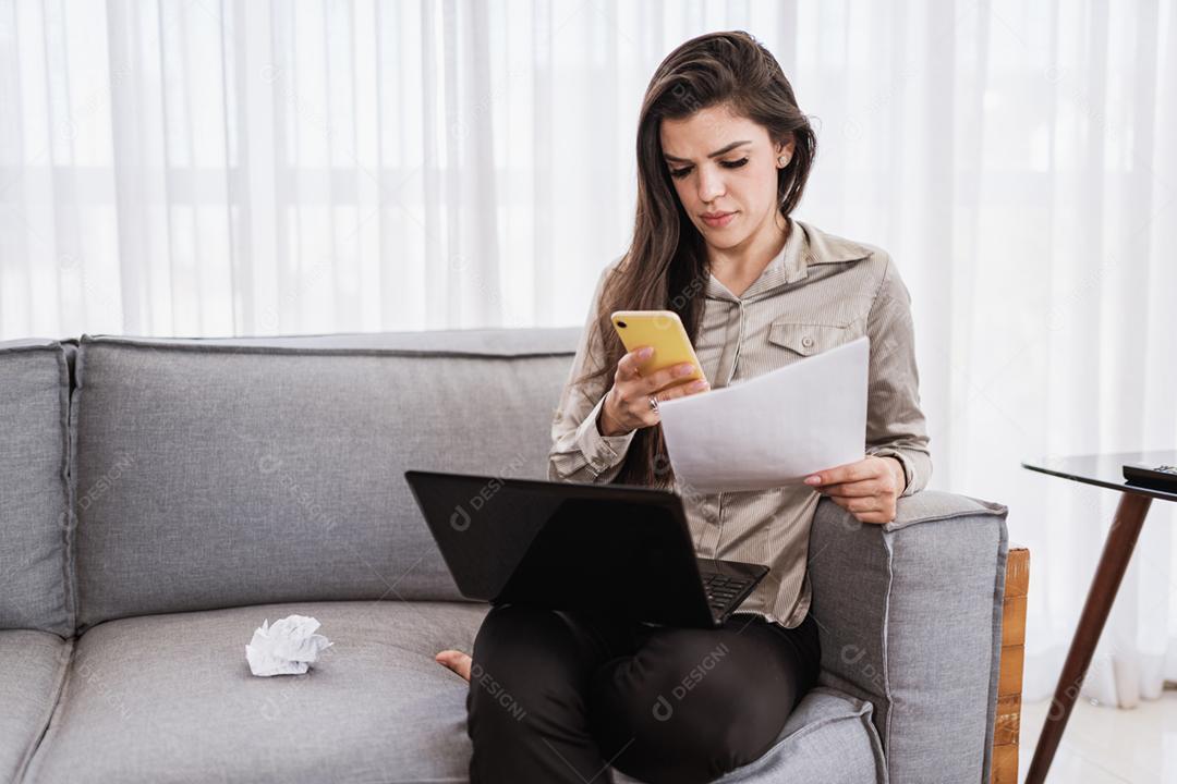 Jovem linda brasileira usando smartphone e trabalhando com laptop enquanto está sentado no sofá