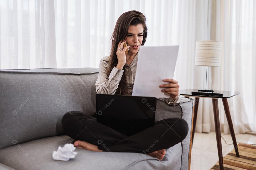 Jovem linda brasileira usando smartphone e trabalhando com laptop enquanto está sentado no sofá