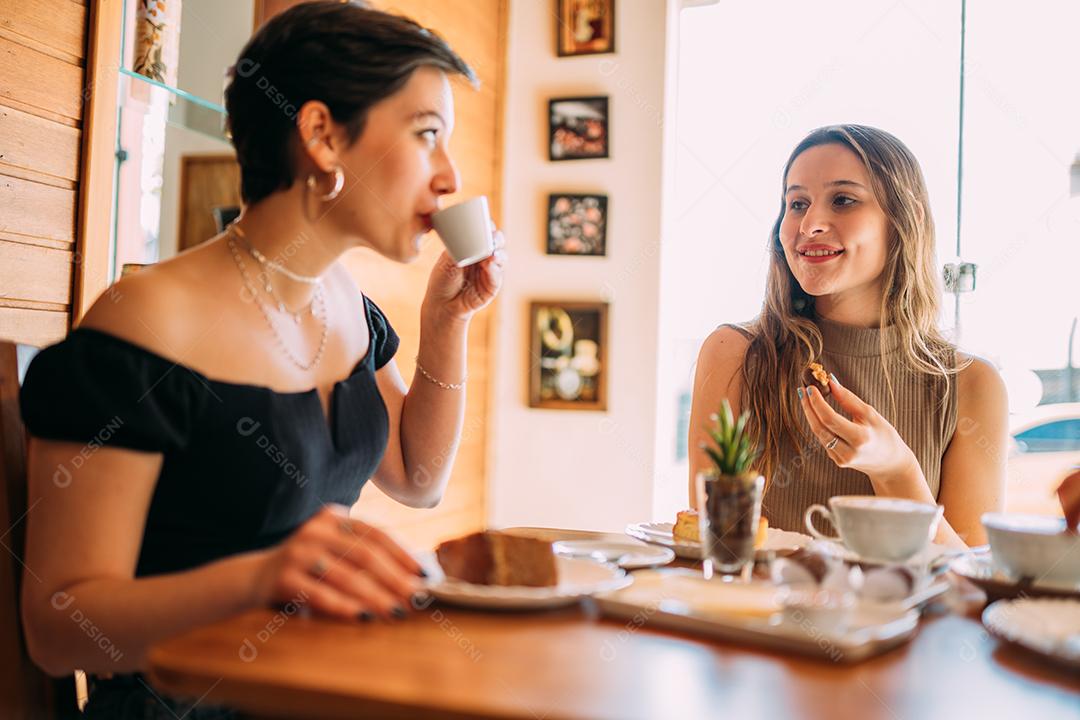 Jovens latinas sentadas em café, tomando café com bolos